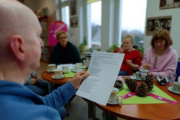 Mieszkaniec czyta tekst z kartki inni siedzą przy stolikach w bibliotece.