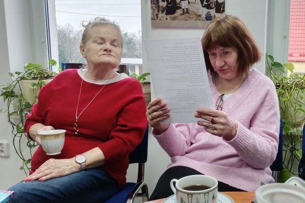 Mieszkaniec czyta tekst z kartki inna mieszkanka siedzi przy stoliku i słucha czytanego tekstu  w bibliotece.