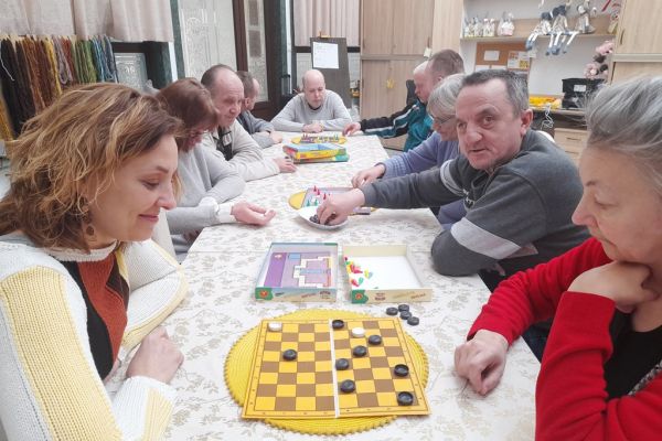 Mieszkańcy siedzą przy długim stole i drużynowo graja w chińczyka.