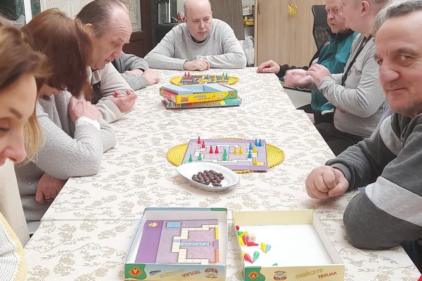 Mieszkańcy siedzą przy długim stole i drużynowo graja w chińczyka.