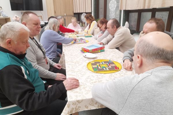 Mieszkańcy siedzą przy długim stole i drużynowo graja w chińczyka.