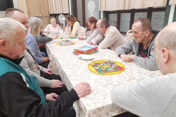 Mieszkańcy siedzą przy długim stole i drużynowo graja w chińczyka.