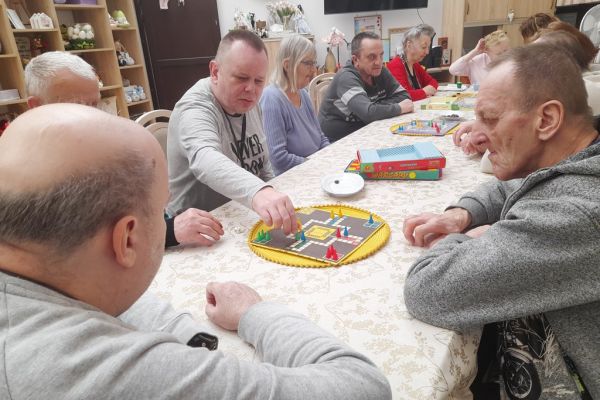 Mieszkańcy siedzą przy długim stole i drużynowo graja w chińczyka.