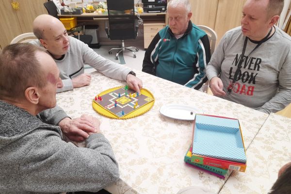 Mieszkańcy siedzą przy długim stole i drużynowo graja w chińczyka.