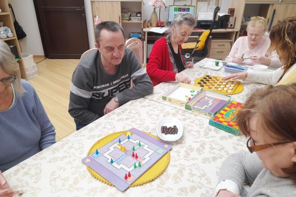 Mieszkańcy siedzą przy długim stole i drużynowo graja w chińczyka.