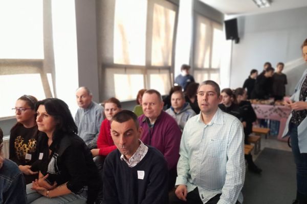 Mieszkańcy siedza w sali szkolnej i oglądają koncert.