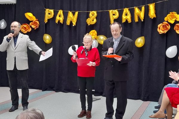 Troje prowadzących uroczystość przemawia do mikrofonów stojąc przed udekorowana kotarą z napisem Pan i Pani.