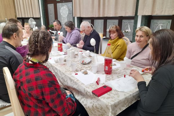 Mieszkanmcy przy długim stole siedzą i wykonują ozdoby świąteczne.