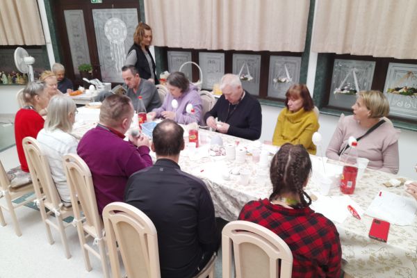 Mieszkanmcy przy długim stole siedzą i wykonują ozdoby świąteczne.