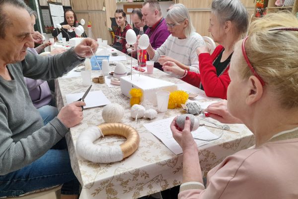 Mieszkanmcy przy długim stole siedzą i wykonują ozdoby świąteczne.