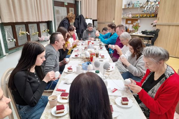 Mieszkańcy wraz z młodzieżą przy długim stole na siedząco owijają jaja sznurkiem.