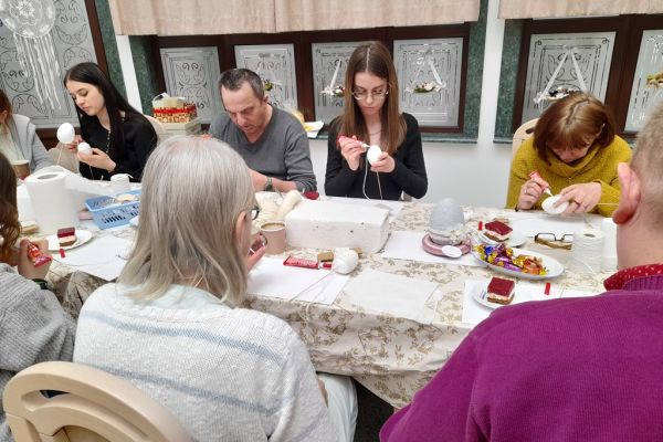 Mieszkańcy wraz z młodzieżą przy długim stole na siedząco owijają jaja sznurkiem.