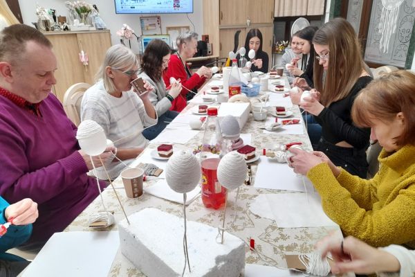 Mieszkańcy wraz z młodzieżą przy długim stole na siedząco owijają jaja sznurkiem.