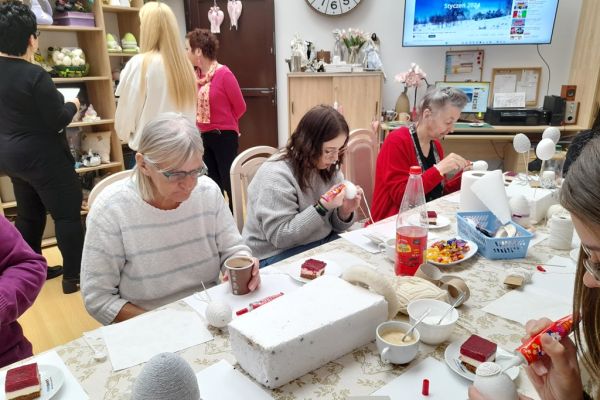 Mieszkańcy wraz z młodzieżą przy długim stole na siedząco owijają jaja sznurkiem.