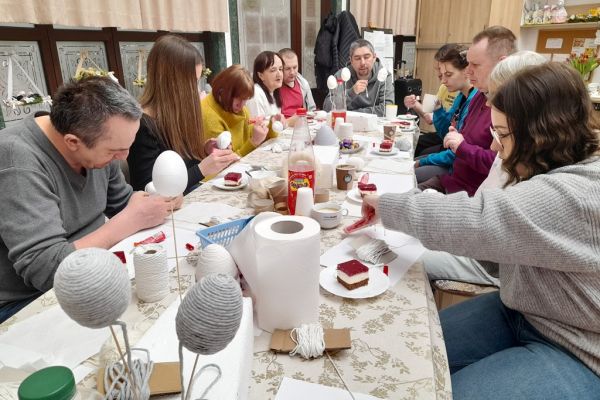 Mieszkańcy wraz z młodzieżą przy długim stole na siedząco owijają jaja sznurkiem.