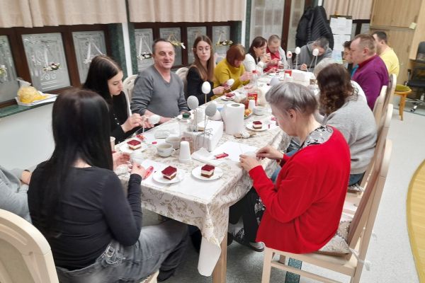 Mieszkańcy wraz z młodzieżą przy długim stole na siedząco owijają jaja sznurkiem.