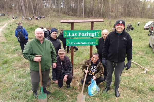 Sześcioro mieszkańców stoi przy tablicy z napisem las posługi.