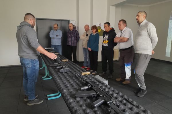 Mieszkańcy na strrzelnicy wirtualnej słuchają wykłady pana o broni stojąc wzdłuż stołu.