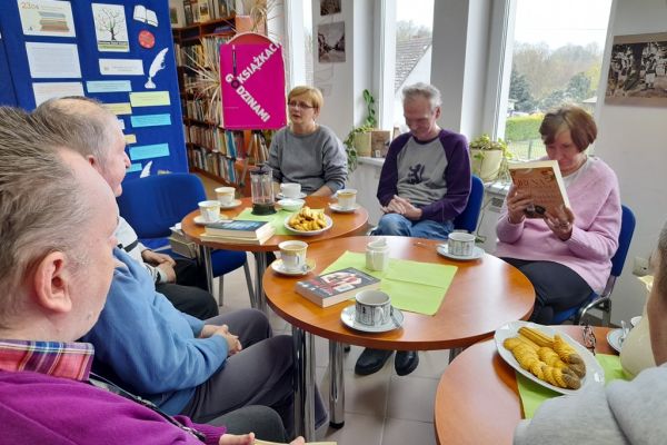 Kilkoro mieszkańców w bibliotece rozmawia o książkach siedząc przy stolikach.