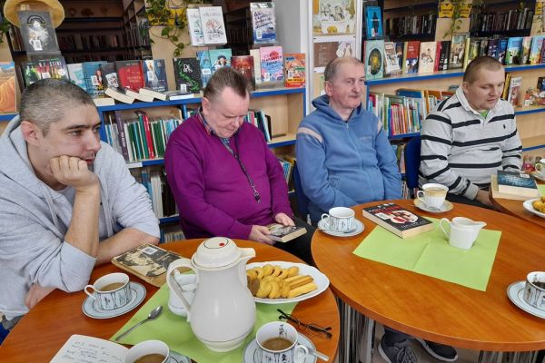 Kilkoro mieszkańców w bibliotece rozmawia o książkach siedząc przy stolikach.