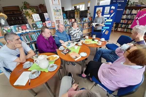 Kilkoro mieszkańców w bibliotece rozmawia o książkach siedząc przy stolikach.