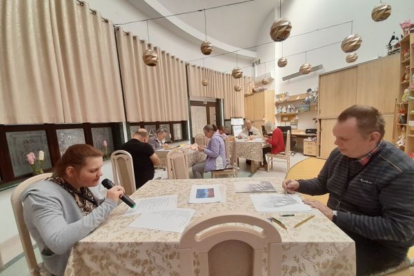 Mieszkańcy wykonują różne rzeczy w sali terapii zajęciowej siedząc przy stolikach.