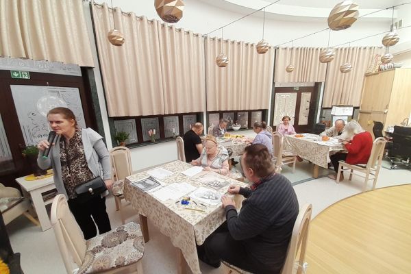 Mieszkańcy wykonują różne rzeczy w sali terapii zajęciowej siedząc przy stolikach.