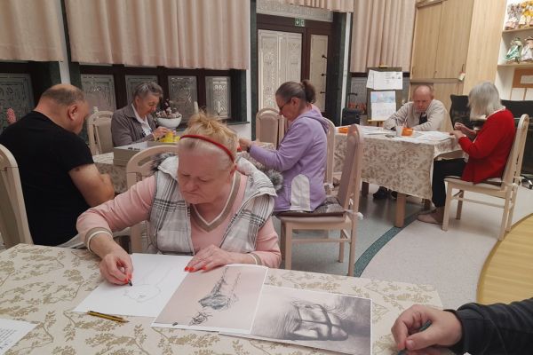 Mieszkańcy wykonują różne rzeczy w sali terapii zajęciowej siedząc przy stolikach.