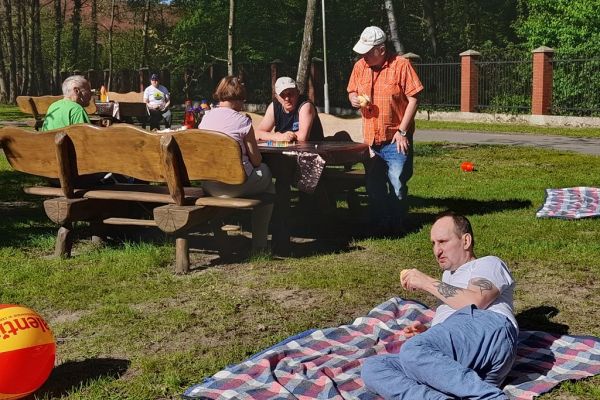 Mieszkańcy siedza na ławeczkach i leżą na kocu na świeżym powietrzu.