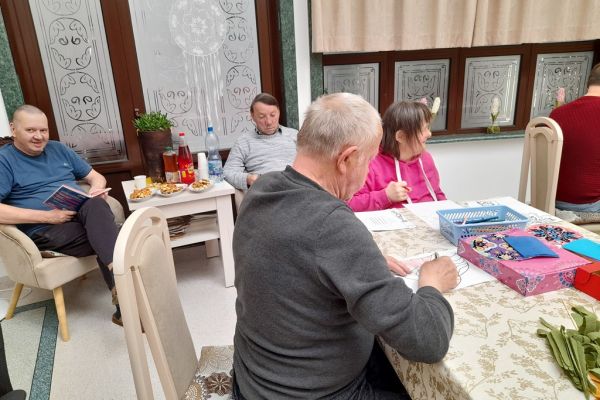 Mieszkańcy w sali terapii siedzą przy stolikach i grają lub rysują lub wykonują inne czynności.