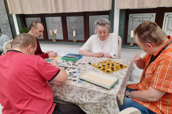 Mieszkańcy w sali terapii siedzą przy stolikach i grają lub rysują lub wykonują inne czynności.