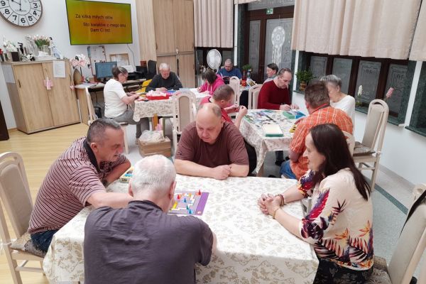 Mieszkańcy w sali terapii siedzą przy stolikach i grają lub rysują lub wykonują inne czynności.