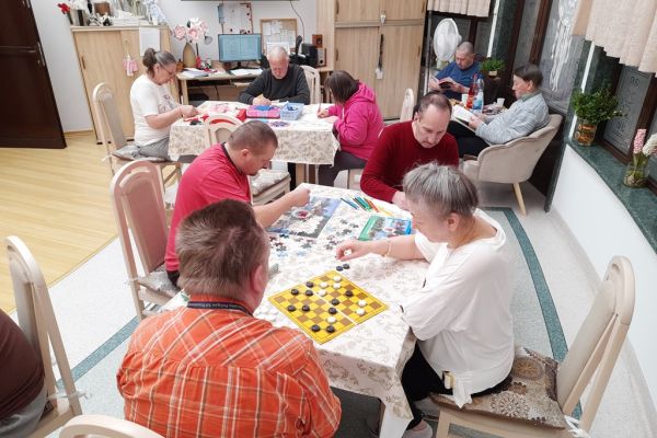 Mieszkańcy w sali terapii siedzą przy stolikach i grają lub rysują lub wykonują inne czynności.