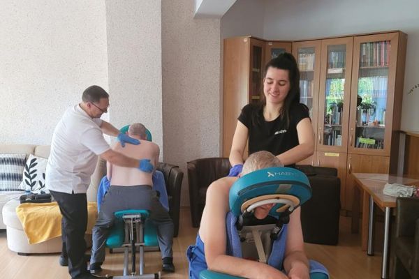 Kilkoro mieszkańców na dużej sali w domu mają wykonywany masaż na leżąco przez ucznów szkoły.