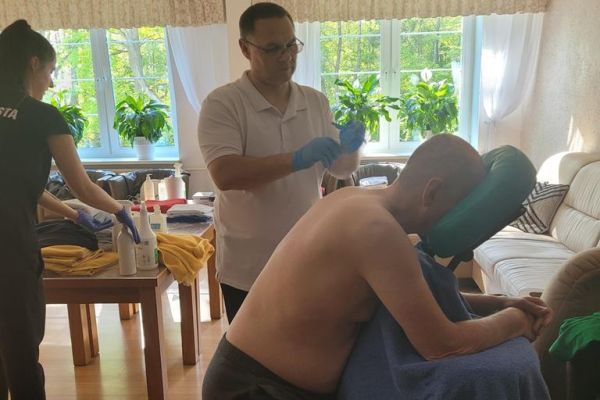 Kilkoro mieszkańców na dużej sali w domu mają wykonywany masaż na leżąco lub siedząco  przez ucznów szkoły.