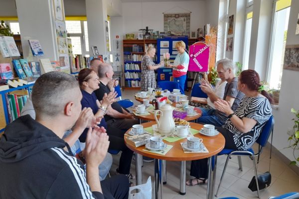 Mieszkańcy na spotkaniu siedza przy okrągłych stolikach i czytają wybrane teksty po kolei.