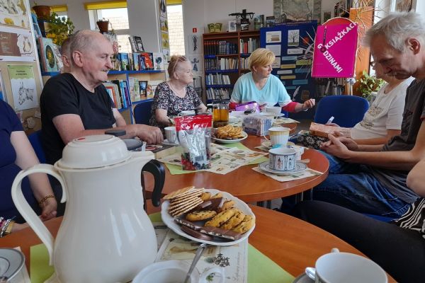 Mieszkańcy na spotkaniu siedza przy okrągłych stolikach i czytają wybrane teksty po kolei.