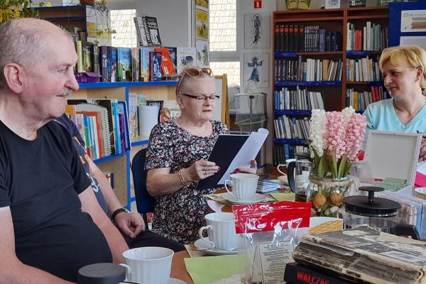 Mieszkańcy na spotkaniu siedza przy okrągłych stolikach i czytają wybrane teksty po kolei.