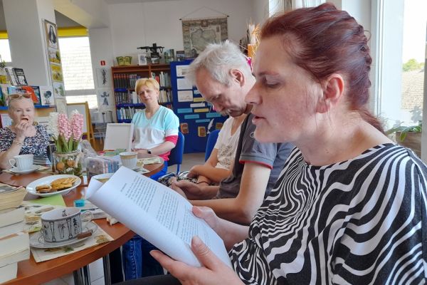 Mieszkańcy na spotkaniu siedza przy okrągłych stolikach i czytają wybrane teksty po kolei.