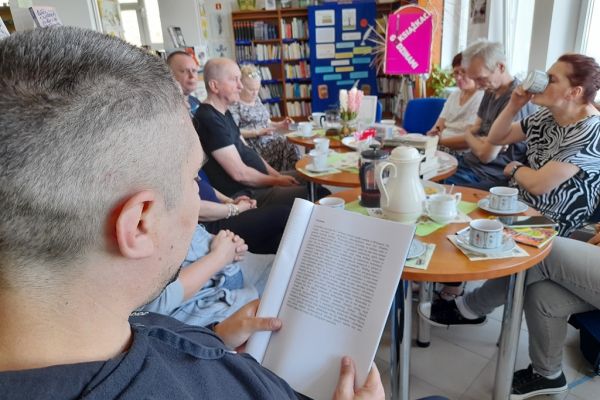 Mieszkańcy na spotkaniu siedza przy okrągłych stolikach i czytają wybrane teksty po kolei.