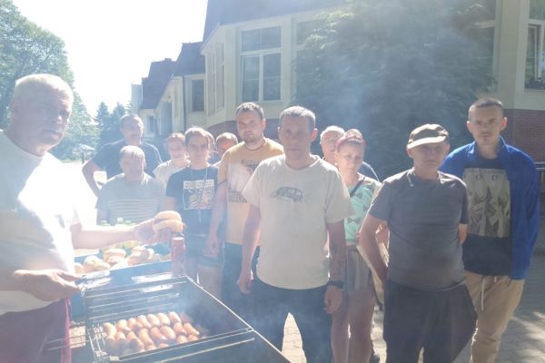 Mieszkańcy odbierają kiełbaski od kucharza, który stoi przy grillu.