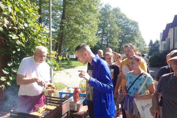 Mieszkańcy odbierają kiełbaski od kucharza, który stoi przy grillu.
