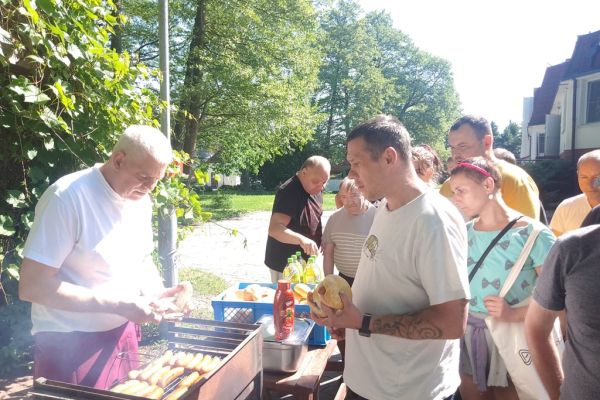 Mieszkańcy odbierają kiełbaski od kucharza, który stoi przy grillu.
