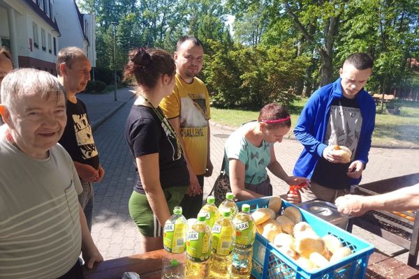 Mieszkańcy odbierają kiełbaski od kucharza, który stoi przy grillu.