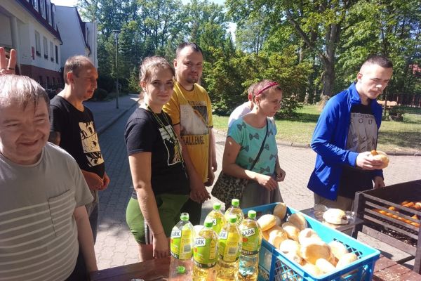 Mieszkańcy odbierają kiełbaski od kucharza, który stoi przy grillu.