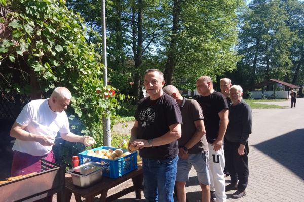 Mieszkańcy odbierają kiełbaski od kucharza, który stoi przy grillu.