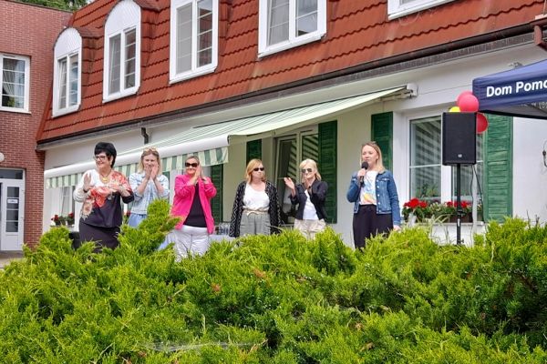 Władze placówek witają uczestników pikniku na stojąco na tle domu.