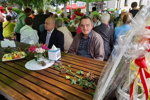 Dwoje mieszkańców pod namiotem przy stoliku w tle inni uczetnicy uroczystości.