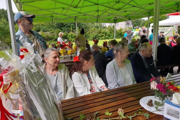 Troje mieszkańców pod namiotem przy stoliku w tle inni uczetnicy uroczystości.