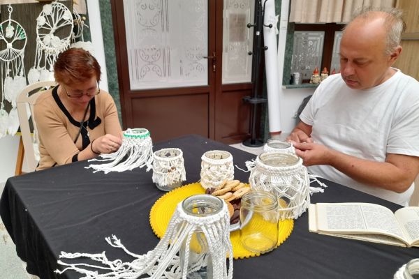 Mieszkańcy w sali terapi siedząc przy stolikach pletą makramę ze sznurka.
